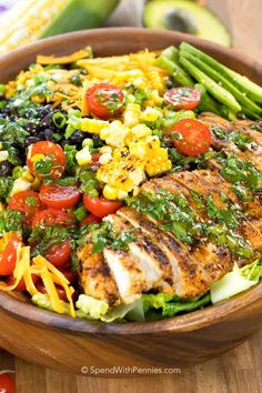 a wooden bowl filled with salad and chicken on top of a table next to corn