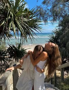 two young women hugging each other on the steps by the beach with palm trees in the background