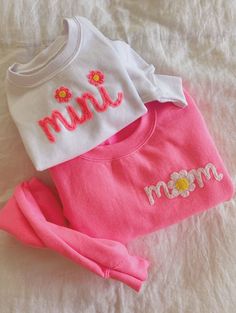 three baby clothes laying on top of a white bed next to each other, one with the word mom written on it