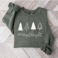 a green sweatshirt that says merry and bright with white trees on the front, sitting on a wood floor