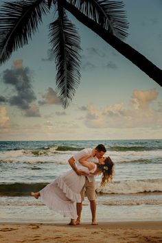 a man and woman kissing on the beach under a palm tree in front of the ocean