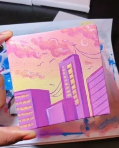 someone is holding up a painting with buildings in the background and pink clouds above them