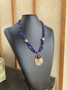 Featuring stones that are associated with wisdom, insight, spirituality, and communication, this is made with gorgeous deep royal blue to navy blue grade A Lapis in various shapes and sizes, which contrast strikingly with green semi precious turquoise, rich red Czech glass and white-heart beads, and brass spacers. The two strands gather at Tibetan brass 15mm coin beads inlaid with red coral and turquoise howlite as well as other brass beads. A 53x37mm plated antique gold large tribal style medal Bohemian Lapis Lazuli Beaded Necklaces As Gift, Bohemian Lapis Lazuli Beaded Necklaces For Gift, Bohemian Lapis Lazuli Beaded Necklace For Gift, Colorful Beads Lapis Lazuli Necklace Gift, Blue Lapis Lazuli Jewelry For Festivals, Handmade Blue Lapis Lazuli Necklace, Handmade Blue Medallion Necklace, Handmade Bohemian Sapphire Necklace, Blue Lapis Lazuli Necklace With Colorful Beads