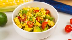 a white bowl filled with corn and vegetables next to a green tomato, an ear of corn on the cob