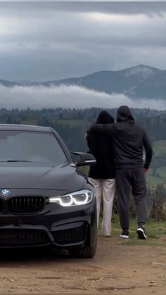 two people standing next to a black car on top of a dirt road with mountains in the background