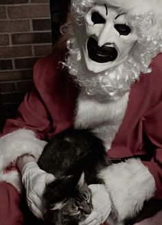 a santa claus holding a cat in his lap while wearing a mask and gloves on it's head