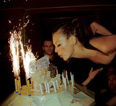 a woman blowing out candles on a cake with her arms in the air as people look on