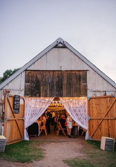 an old barn with white curtains and people sitting at tables in the doorway, surrounded by green grass