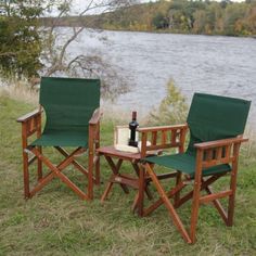 two wooden chairs and a table with a bottle of wine on the grass near water