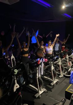 a group of people riding stationary exercise bikes in a dark room with their arms up