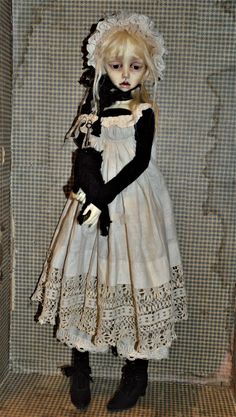 a doll is dressed in an old fashion dress and holding a black teddy bear while standing next to a wall