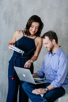 a man and woman sitting on a stool looking at an open laptop computer, both smiling