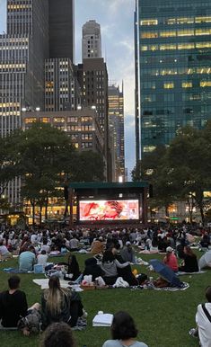 many people are sitting in the grass watching a movie on a big screen at dusk