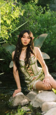 a woman in a fairy costume sitting on rocks next to a stream with her legs crossed