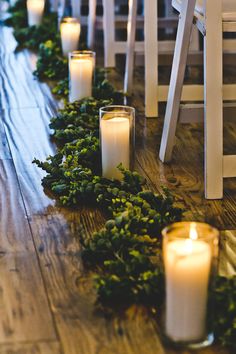 candles are lined up on the floor in front of white chairs and greenery garland