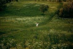 a woman walking through a lush green field next to trees and wildflowers on a hillside