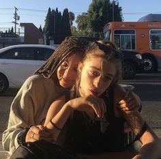 two young women sitting on the ground in front of a street with cars behind them