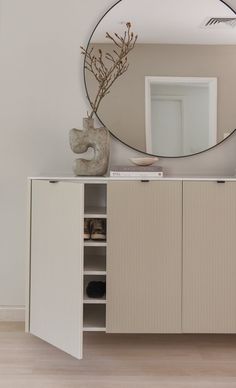 a white cabinet with a round mirror above it and a vase in front of it