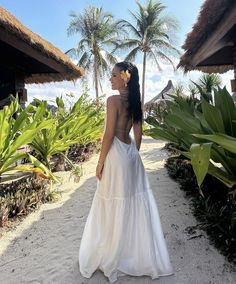 a woman in a white dress is standing on the sand near some palm trees and bushes