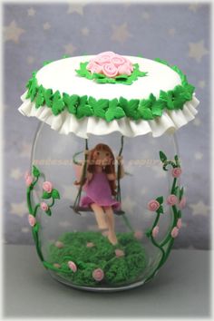 a doll in a glass jar with flowers and grass on the bottom, holding a swing