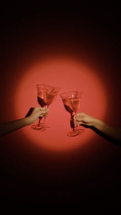 two hands holding up wine glasses in front of a red background with the light shining on them