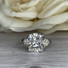 a white rose sitting next to a ring on top of a wooden table with two diamonds