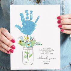 a woman holding up a card with a handprint on it and flowers in a mason jar