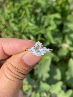 a person holding an engagement ring in their left hand, with green plants behind them