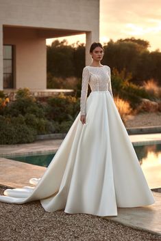 a woman standing in front of a pool wearing a long sleeved wedding dress with an open back