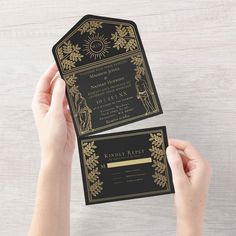 two hands holding up black and gold wedding cards