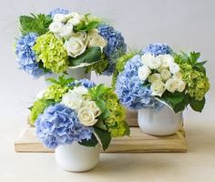 three white vases filled with blue and green flowers on a wooden stand in front of a wall