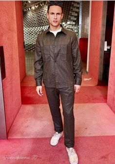 a man standing in front of a red wall wearing a black shirt and grey pants