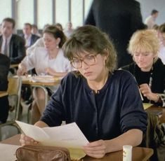 a woman sitting at a desk with papers in front of her and other people behind her