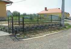 an iron fence is in front of a yellow house and gravel road with rocks on the ground