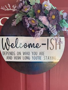 a welcome sign hanging on a red door with purple flowers and greenery in it