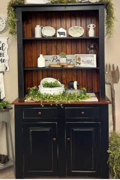 a black china cabinet with green plants on top and white dishes in the bottom shelf