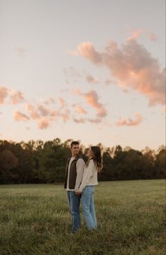 a man and woman standing in the grass under a pink sky with clouds above them