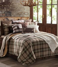 a bed with plaid comforter and pillows in front of a brick wall next to a window