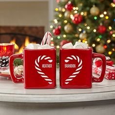 two red coffee mugs sitting on top of a table next to a christmas tree