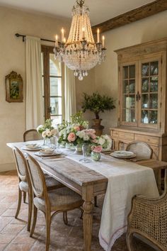 A charming French countryside-inspired dining room featuring a rustic wooden table paired with cane-back chairs. The table is elegantly set with a simple linen tablecloth, delicate floral arrangements in soft pastel hues, and crystal glassware. A stunning crystal chandelier hangs above, casting a warm glow over the room, which is complemented by a vintage wooden cabinet and lush greenery. The terracotta tiled floor and antique decor elements create a cozy and inviting French Country Breakfast Rooms, Vintage Cane Back Dining Chairs, French Country Dinner Table, English Cottage Style Dining Room, French Dining Room Ideas, Italian Country House Interior, English Cottage Dining Room, French Country Table Decor, Cottagecore Dining Room