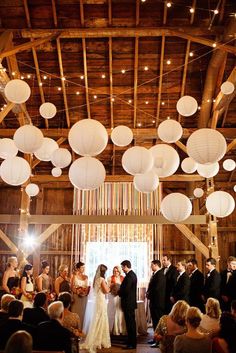 a couple getting married under paper lanterns at their wedding ceremony in the barn with friends and family