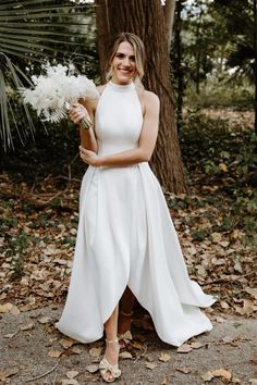 a woman standing in front of a tree holding a bouquet of flowers and wearing a white dress