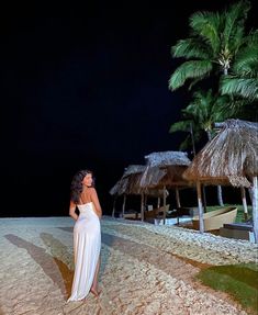 a woman in a white dress standing on the beach at night with palm trees and thatched umbrellas