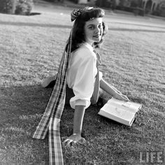 a woman sitting in the grass with a book and scarf around her neck, looking at the camera