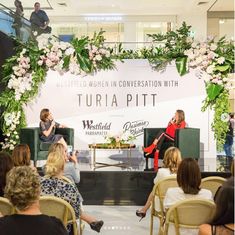 two women sitting on chairs in front of a stage with flowers and greenery around them