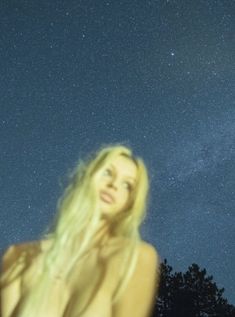 a woman with long blonde hair standing in front of the night sky and looking up at the stars