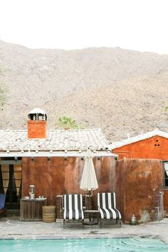 two chairs and an umbrella near a pool in front of a house with mountains behind them