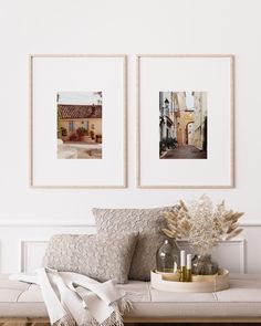 two framed pictures hang on the wall above a bench with pillows and vases next to it