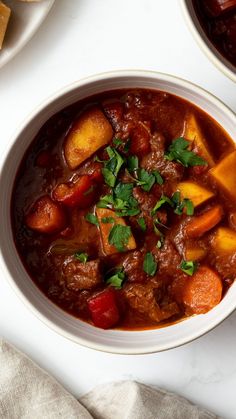 two bowls filled with stew and vegetables on top of a white tablecloth next to bread