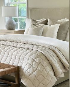 a bed with white comforter and pillows next to a lamp on a table in front of a window